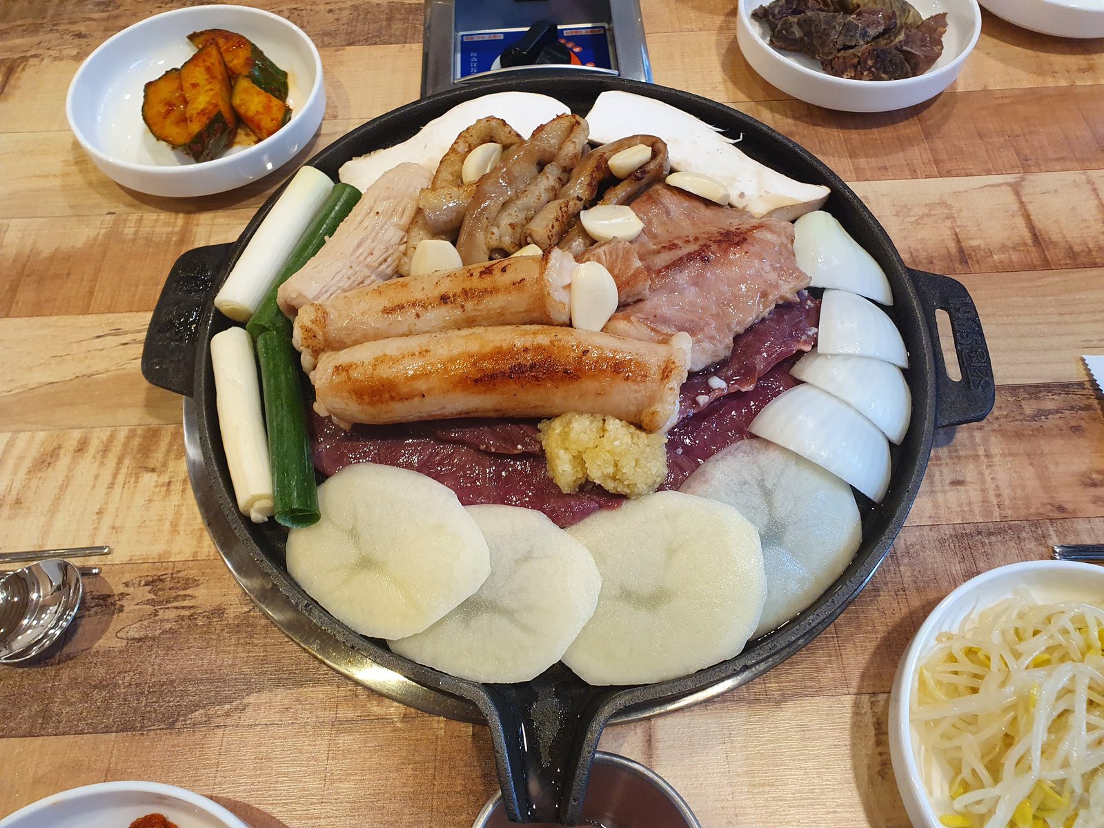 한우소금모듬구이 한 판. 둥근 판 가운데 겉을 노릇하게 익힌 창자 부위와 여러 부위를 모아 통마늘을 올리고, 둘레에 감자·양파·새송이·대파를 둘렀다. 옆에 오이무침과 콩나물무침이 놓여 있다